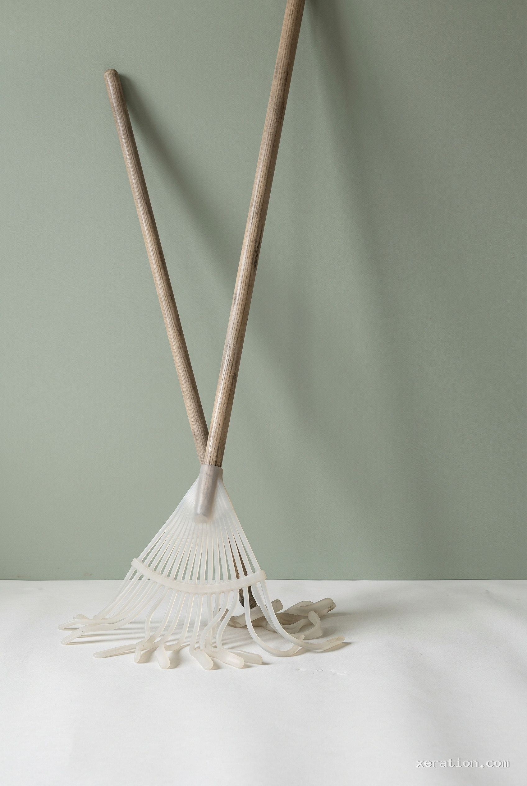 A garden rake leaning against a pale wall, its fan tines replaced by limp translucent silicone strips resting on the floor.